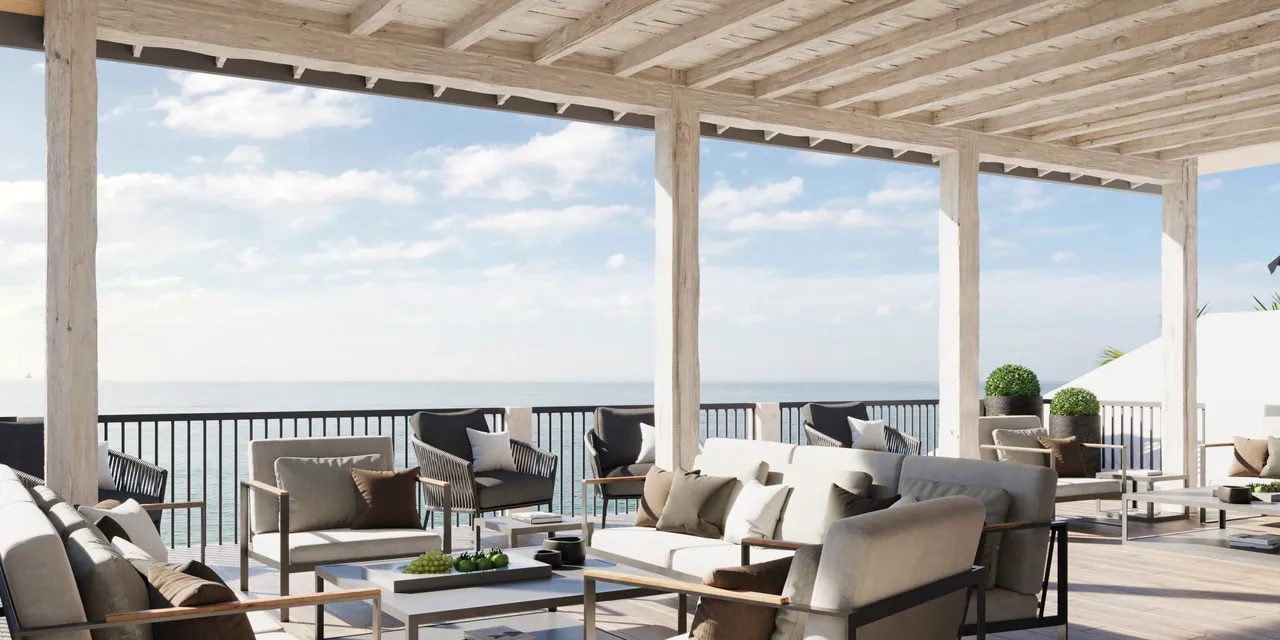 Rendered image of a coastal terrace with modern outdoor seating under a wooden pergola, overlooking a serene ocean view under a bright, partly cloudy sky.